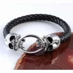 Brazalete bisutería hombre con calaveras acero y cordón piel trenzada negra, sobre fondo gris oscuro.